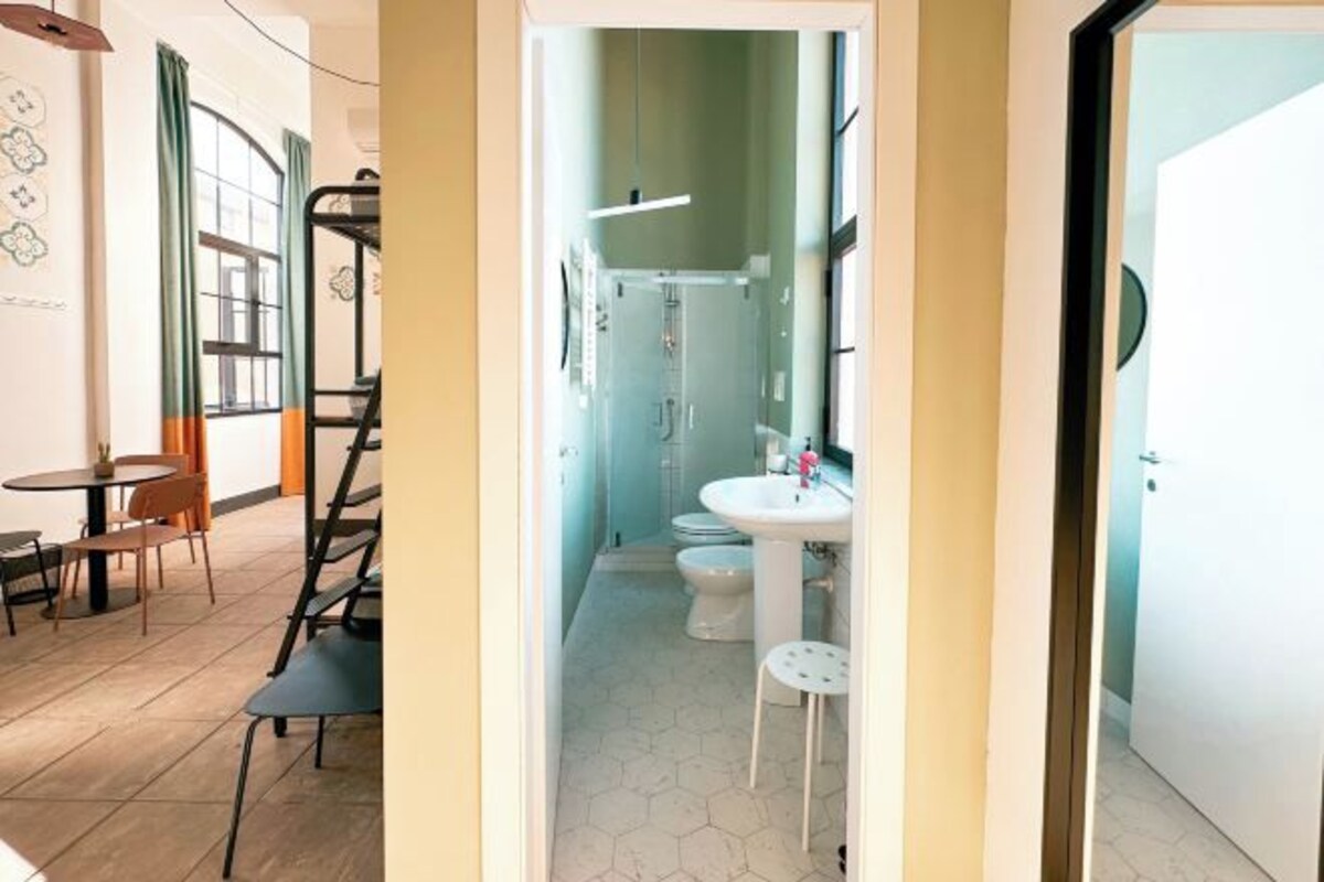 A narrow hallway leading to a bathroom with a toilet, sink, and shower. There is a small table and chairs in the adjacent room.