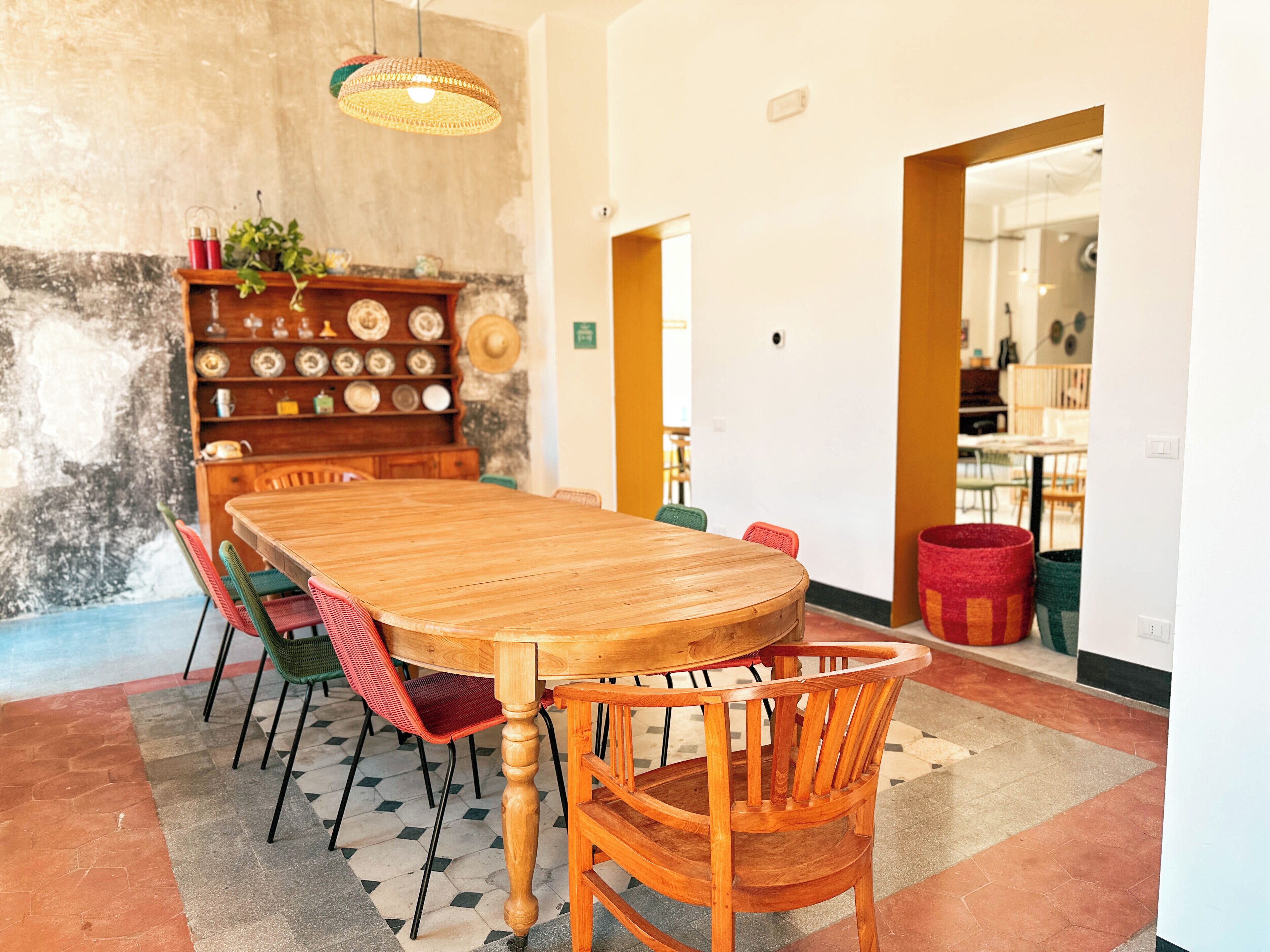 A dining area with a wooden table and chairs, a shelf with plates, and a hanging light fixture.