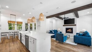 A modern kitchen with white cabinetry, a central island, and pendant lighting. There is a living area with blue sofas and a fireplace. - Cote D' Azur • Private Pool Retreat on 30A • Golf Cart 🛺 (Seagrove Beach)