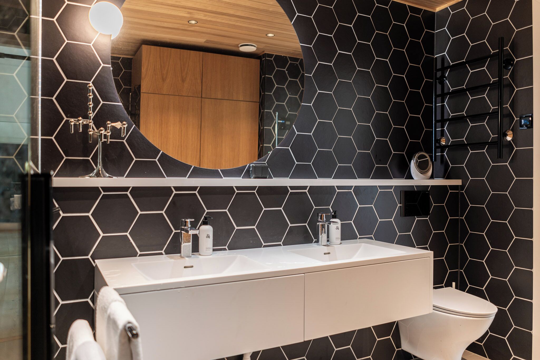A modern bathroom with a black and white hexagonal tile backsplash, a wooden vanity, a double sink, and a toilet.