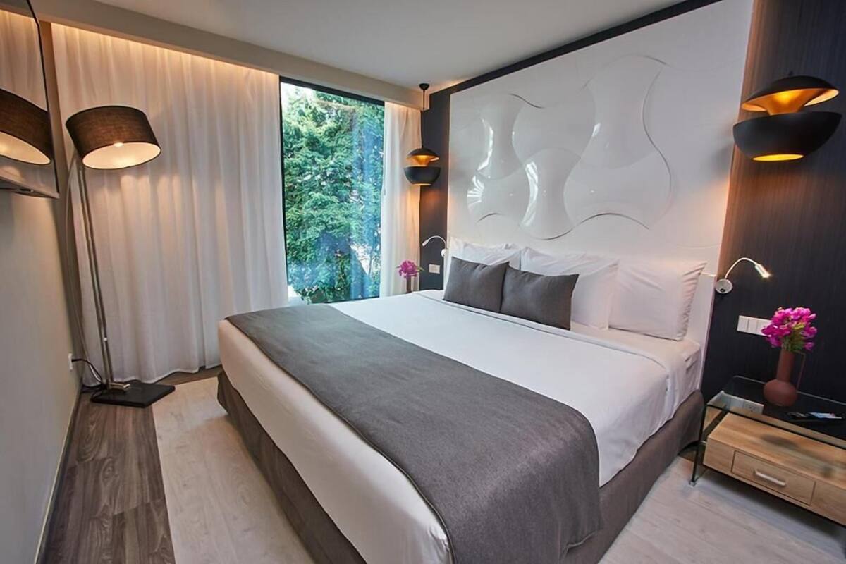 Een moderne slaapkamer met een groot bed, een hoofdbord met een uniek ontwerp, nachtlampjes en uitzicht op groen buiten het raam.