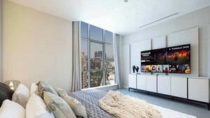 A modern bedroom with a large bed, a view of the city through floor-to-ceiling windows, and a wall-mounted TV displaying a movie poster.