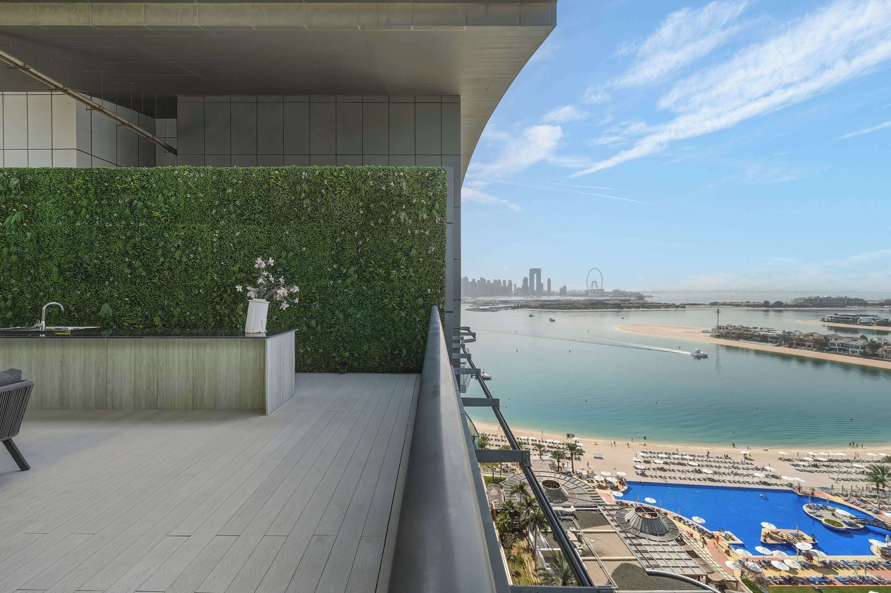 A balcony with a green wall, a pool, and a view of a beach and city skyline.