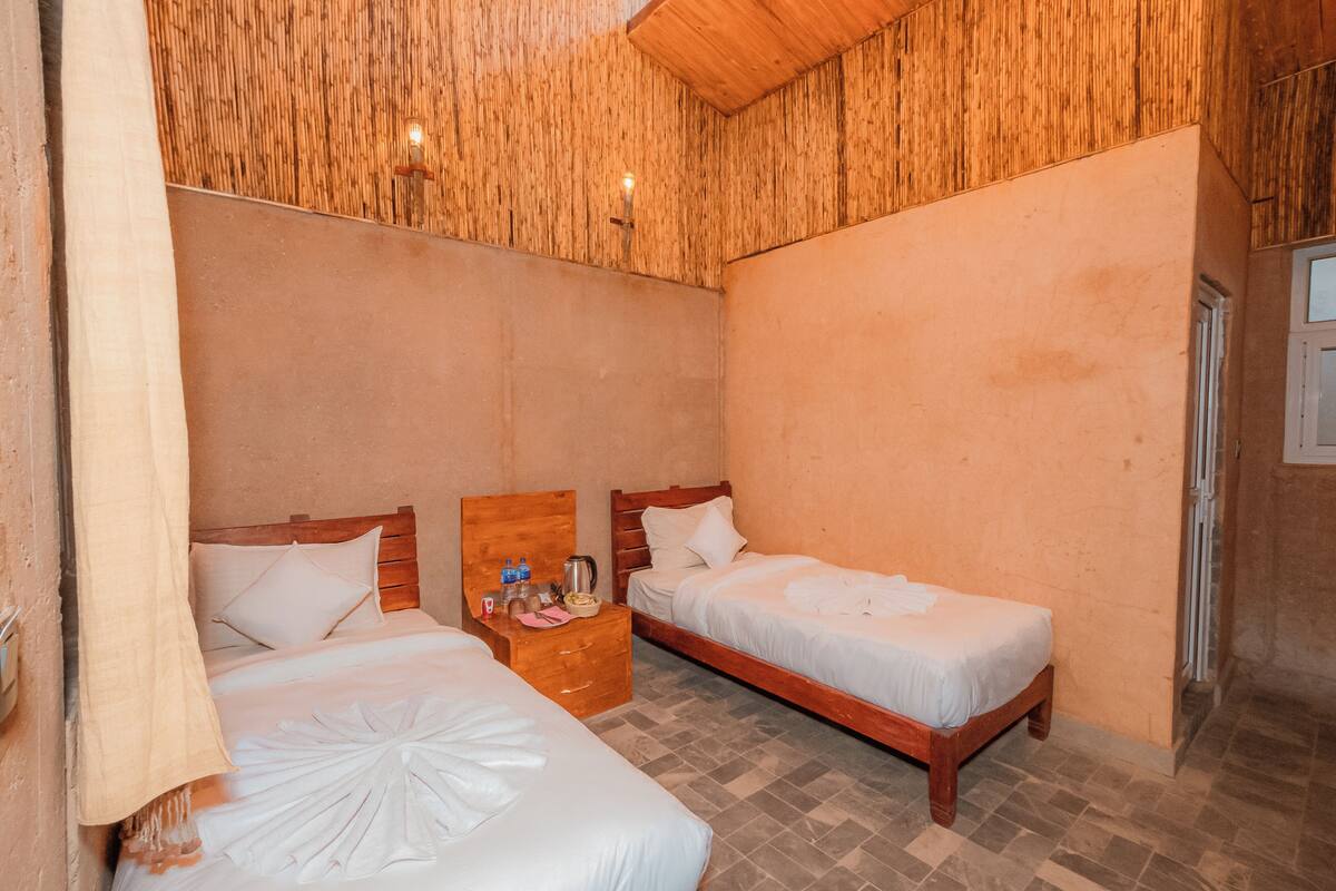 A room with two beds, wooden headboards, a wooden nightstand with a kettle, and a wall-mounted light fixture.