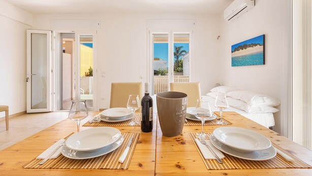 A dining area with a wooden table set for two, white plates, wine glasses, and a bottle of wine. There is a large vase on the table, and a painting of a beach scene on the wall. - 3951 Mono di Annahouse by Barbarhouse (Porto Cesareo)
