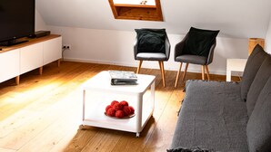 Un salon moderne avec un plafond en pente, un plancher en bois, un canapé gris, deux fauteuils, une table basse blanche sur laquelle se trouve un saladier de fruits, et un téléviseur posé sur un meuble blanc.