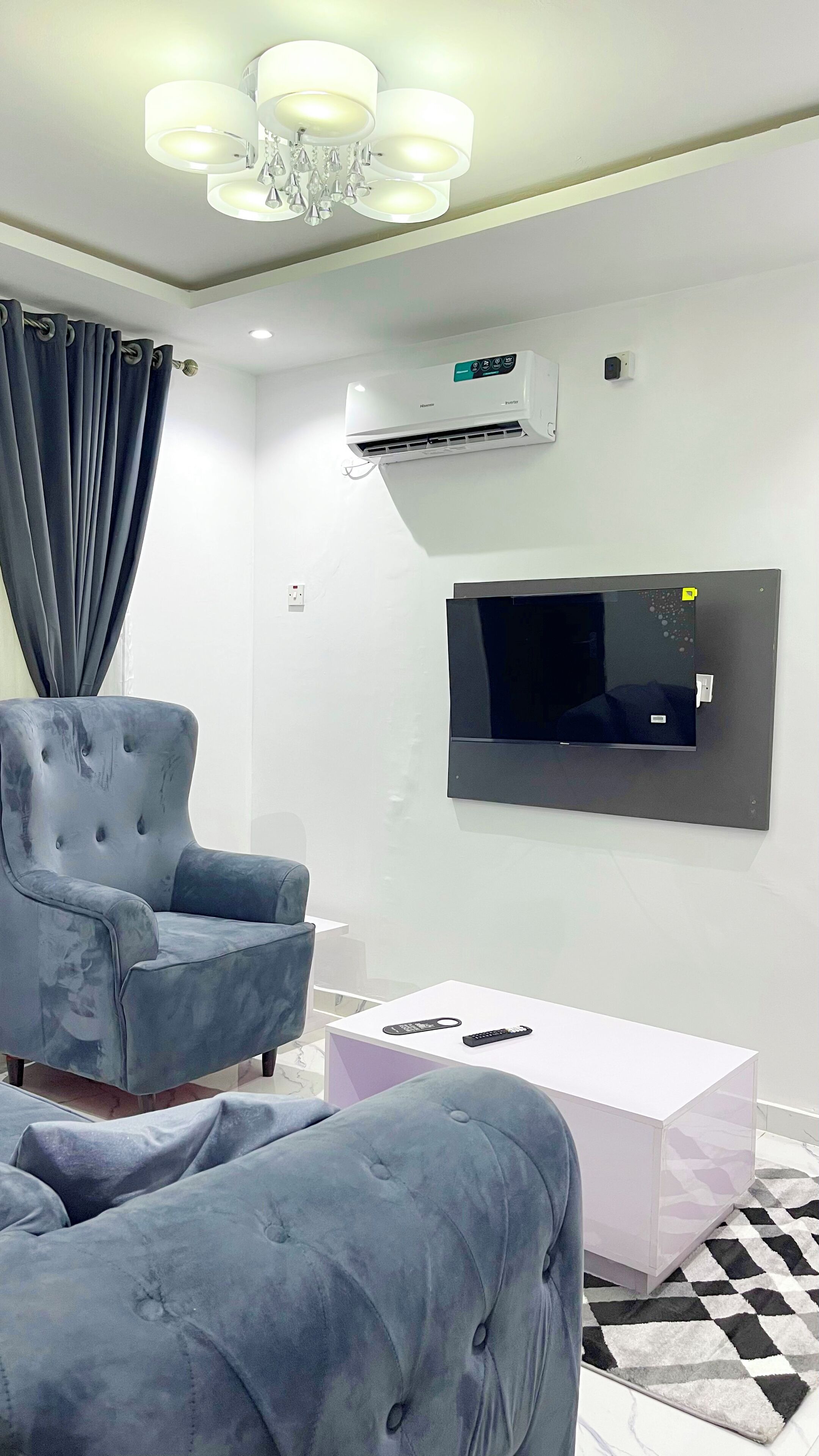 A modern living room with a grey upholstered armchair, a white coffee table, a flat-screen TV mounted on the wall, and a ceiling-mounted air conditioning unit.