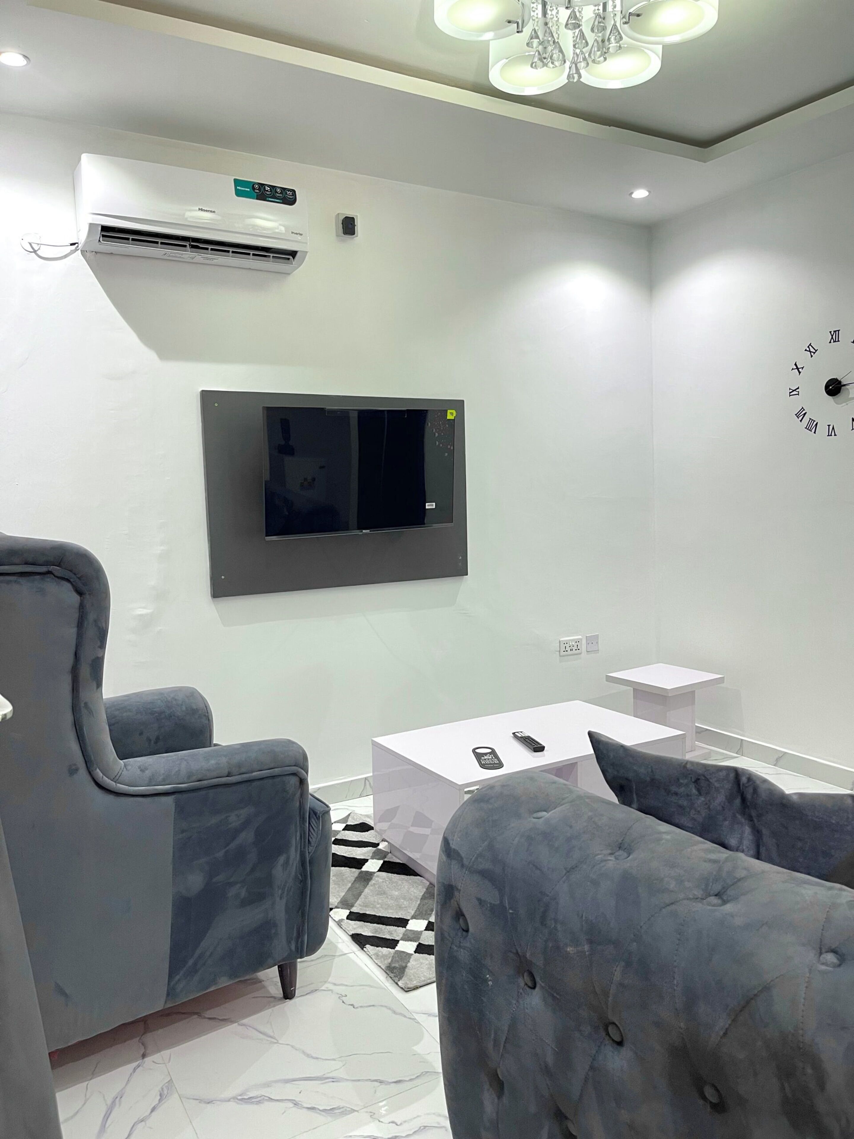 Uma sala de estar moderna com televisão na parede, poltronas revestidas de cinza, uma mesa de centro branca e um tapete estampado.