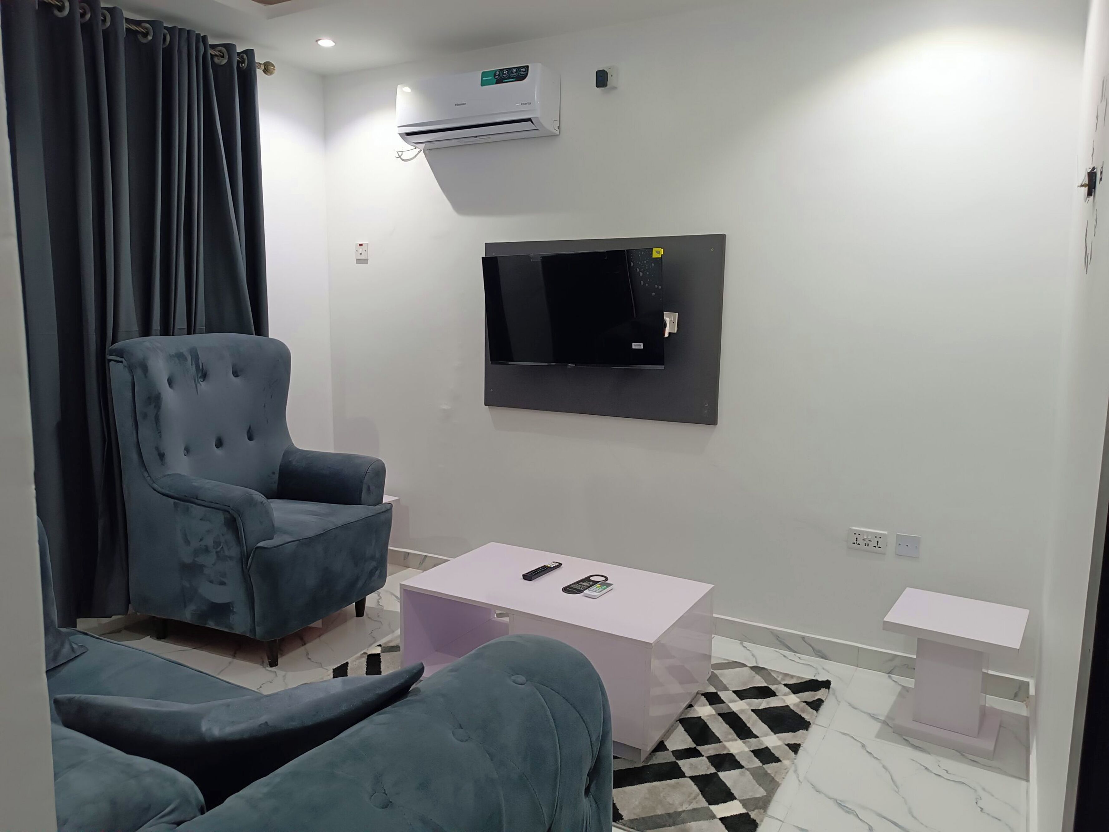 Un salon moderne avec un canapé gris, une petite table basse, un téléviseur à écran plat fixé au mur et un appareil de climatisation.