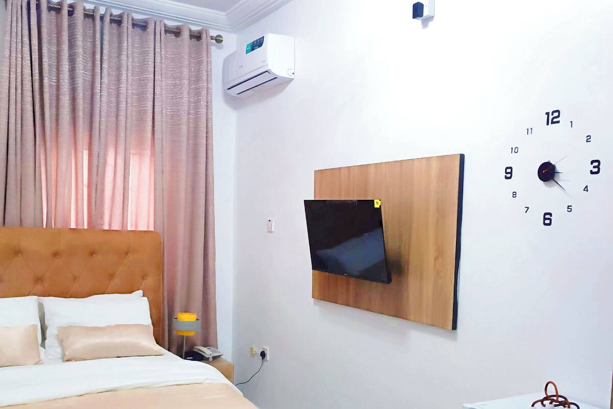 A hotel room with a bed, a TV mounted on the wall, a clock, and a small refrigerator.