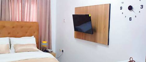 A hotel room with a bed, a TV mounted on the wall, a clock, and a small refrigerator.