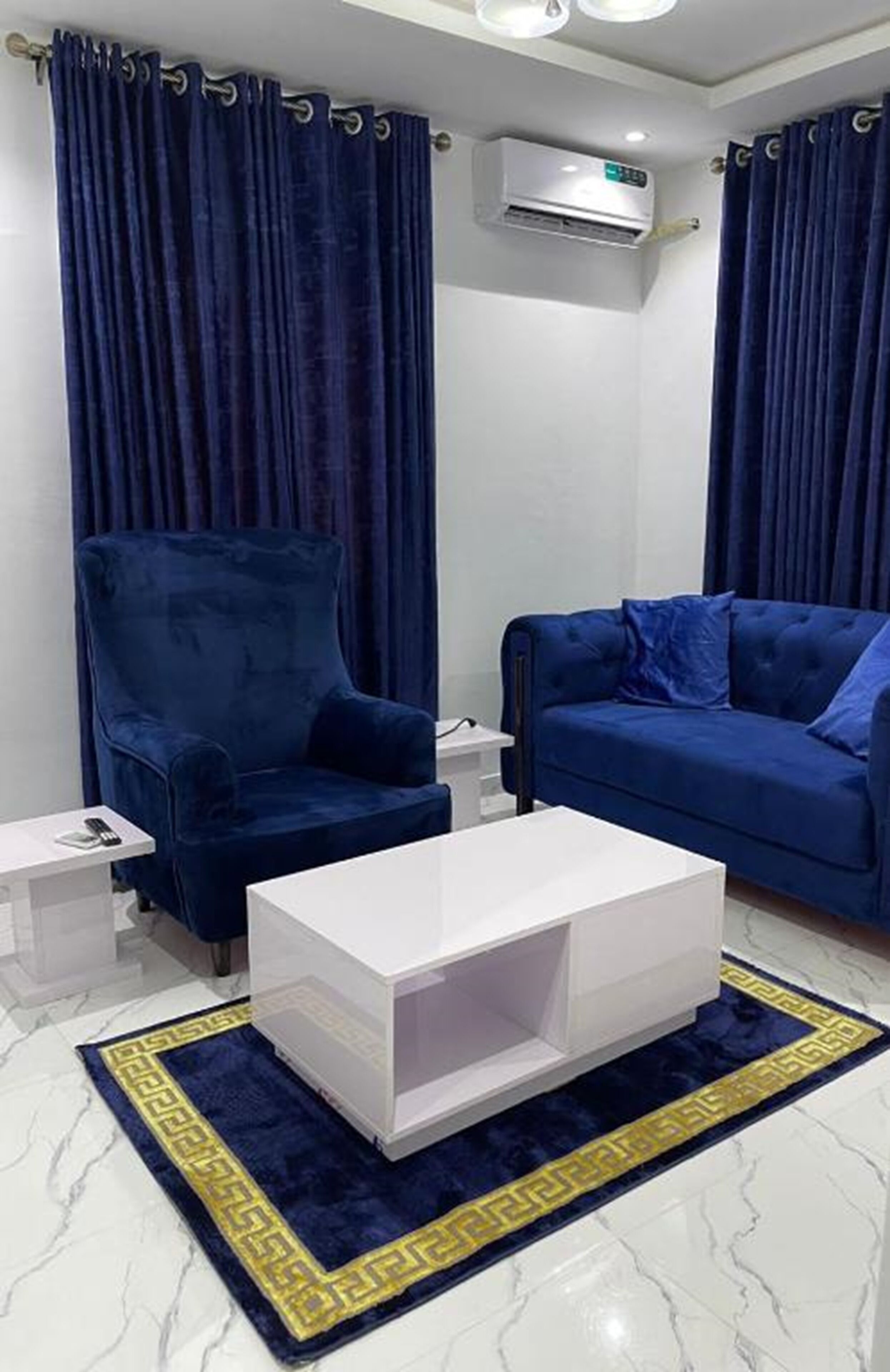 A modern living room with a blue velvet armchair, a blue sofa, and a white coffee table. There is a small white table and a blue rug with a yellow border.