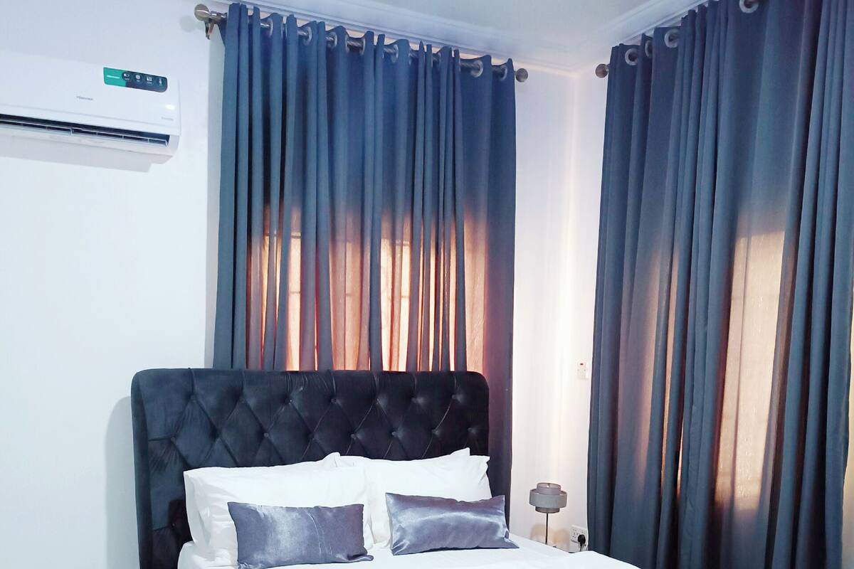 A bedroom with a tufted headboard, a bed with white and dark blue bedding, and blue curtains.