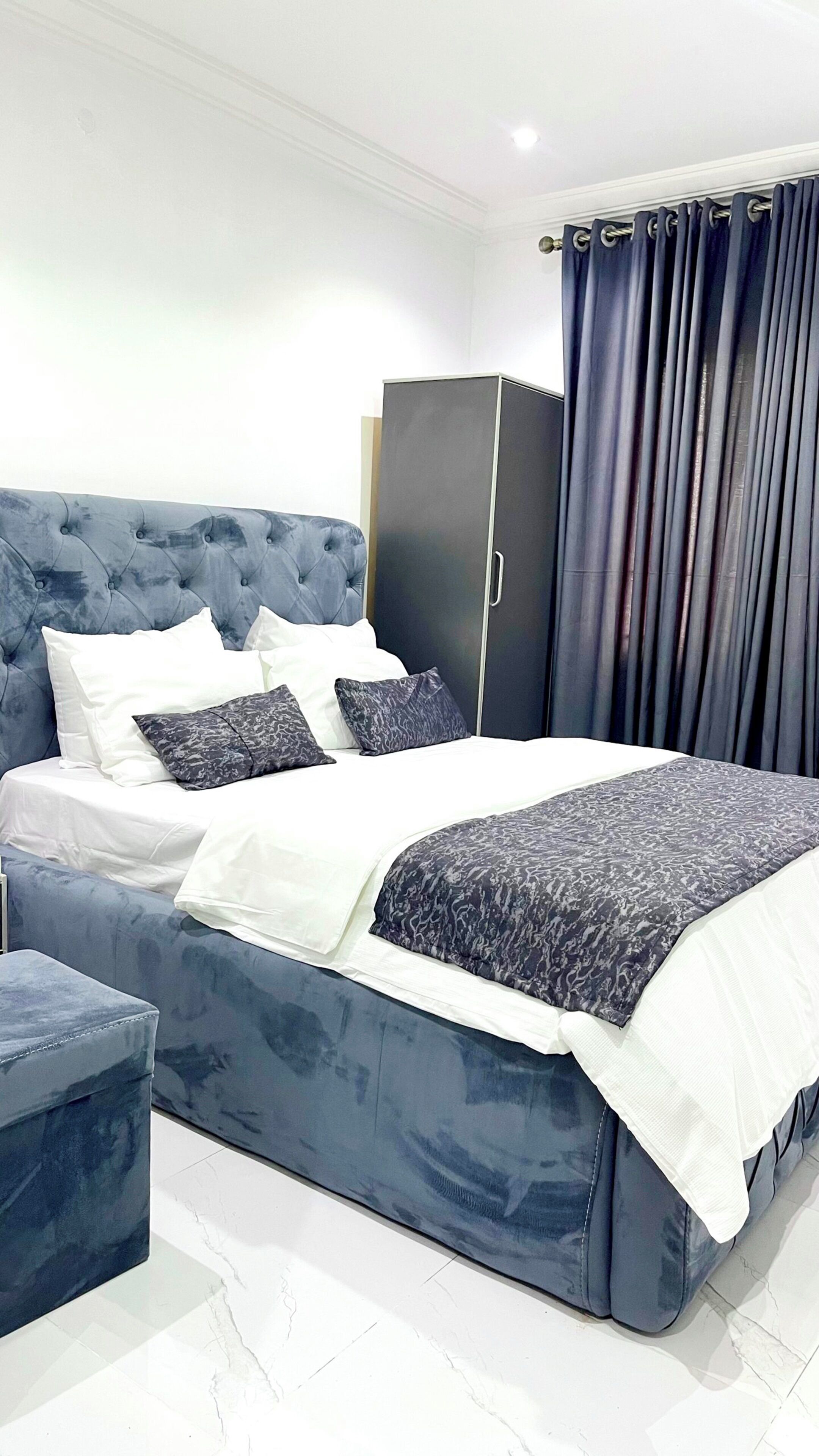 A modern bedroom with a tufted headboard, a large bed with a patterned bedspread, a dark wardrobe, and a matching ottoman.