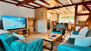 A cozy living room with teal upholstered sofas, a wooden coffee table, and a large flat-screen TV displaying a snowy landscape. - Large Holiday Home for Groups up to 17 Guests with Hot Tub & Playroom (Herbstein)