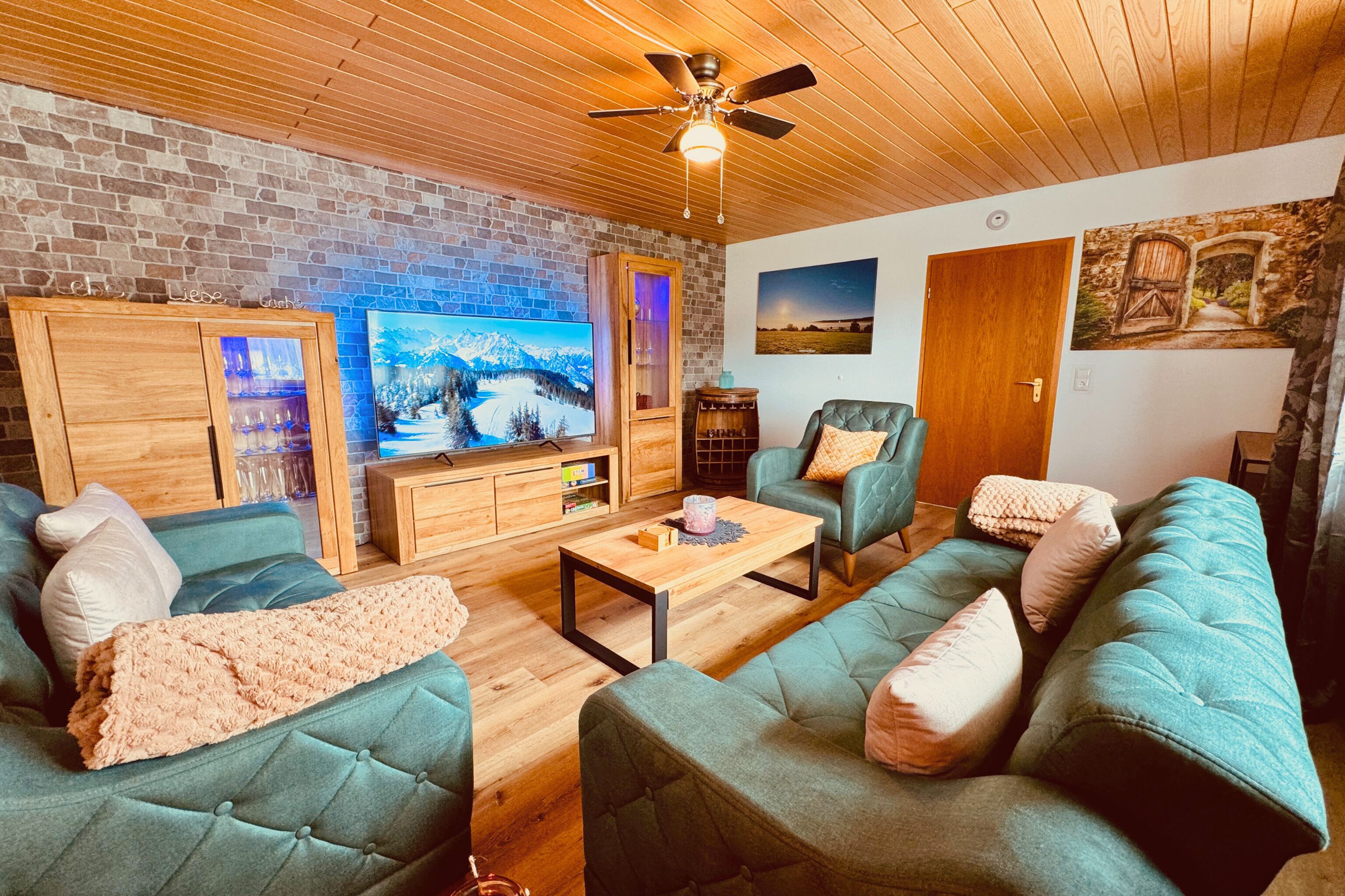 A cozy living room with a brick wall, a wooden ceiling, and a television. There are two green sofas, a wooden coffee table, and a fireplace.