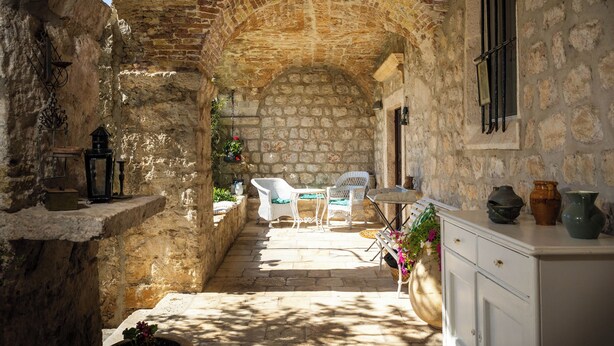 Un pasillo de paredes de piedra con decoración rústica, que incluye una pequeña mesa y sillas, un armario blanco y una ventana con barrotes.