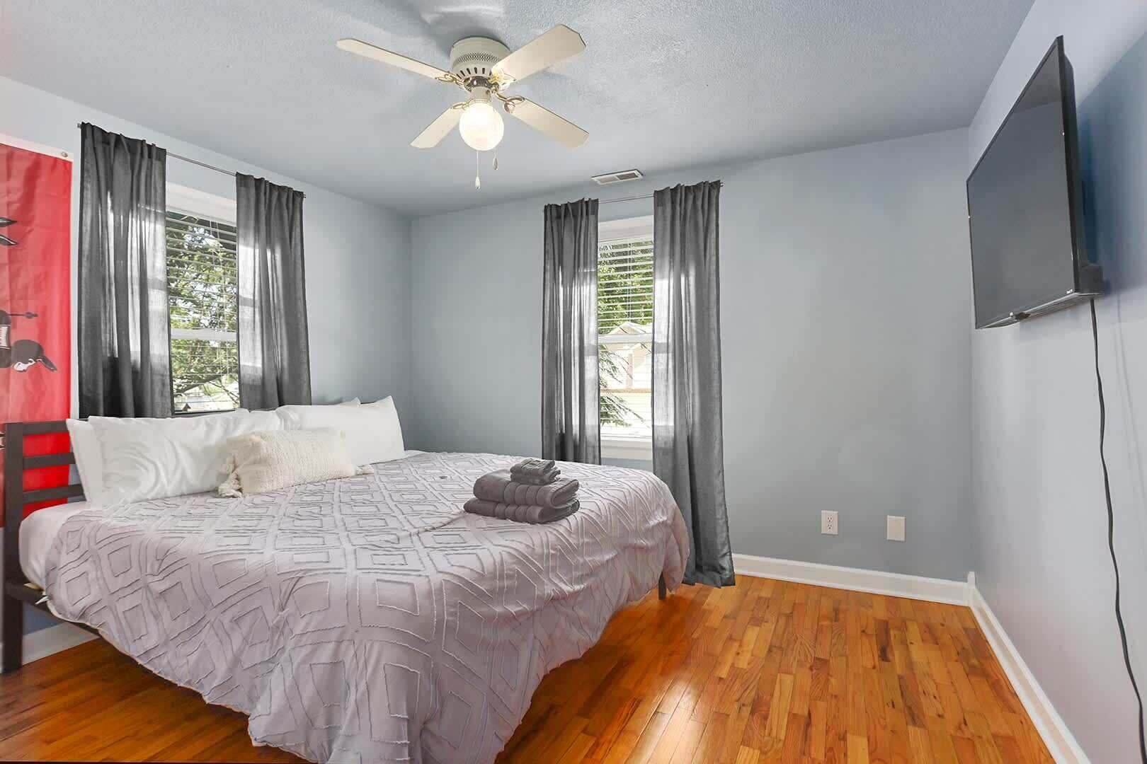 A bedroom with a bed, a ceiling fan, a TV mounted on the wall, and a window with curtains.