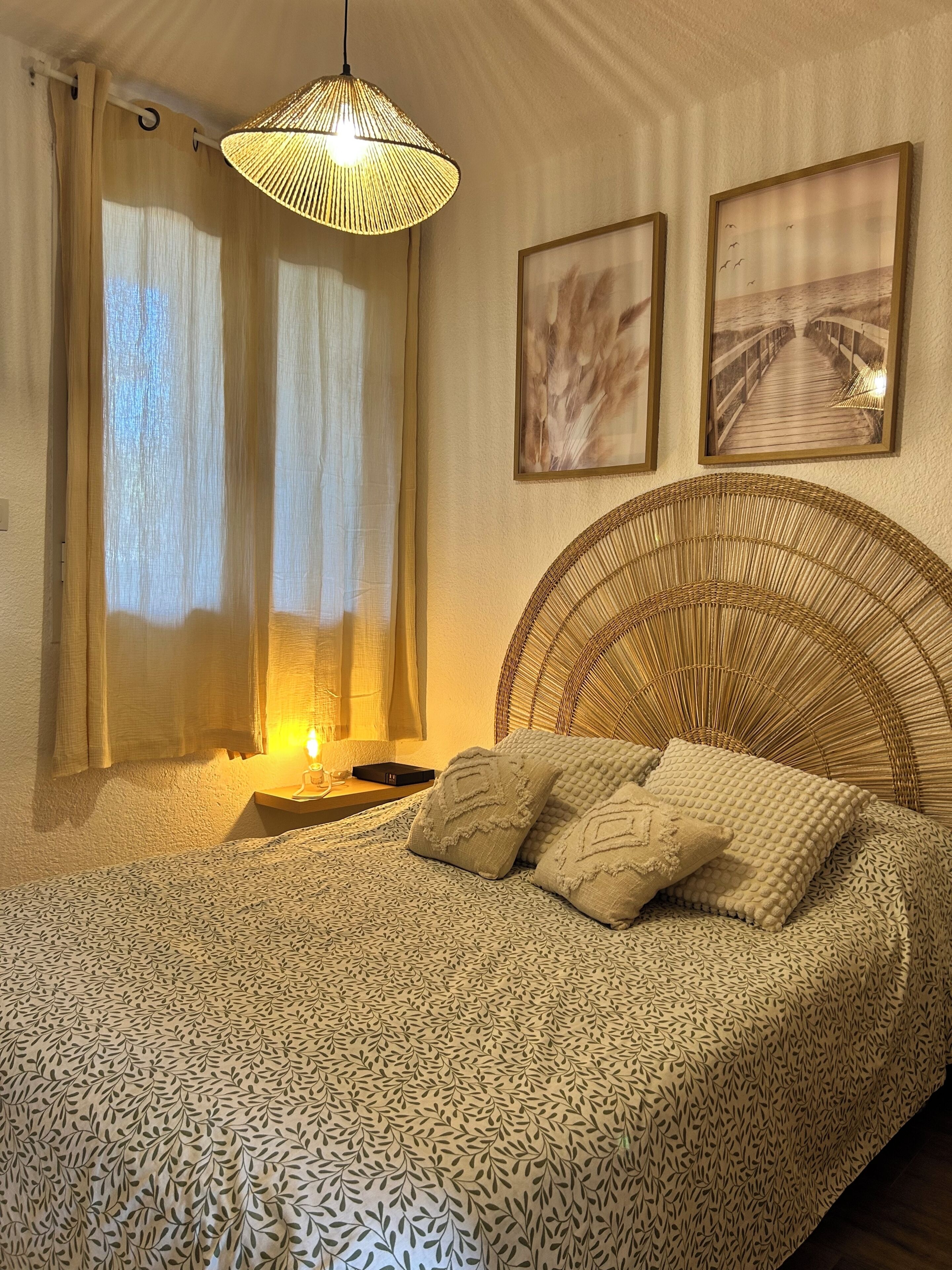 A bedroom with a patterned bedspread, a wicker headboard, a bedside lamp, and two framed pictures on the wall.