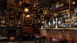 A bar with wooden bar stools, a fireplace, and various bottles and glasses on shelves. - The Olde Ship Inn (Seahouses)