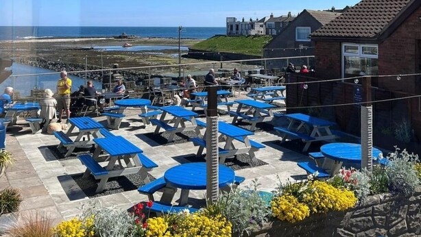 A patio area with blue tables and chairs, overlooking a beach and houses. - The Olde Ship Inn (Seahouses)