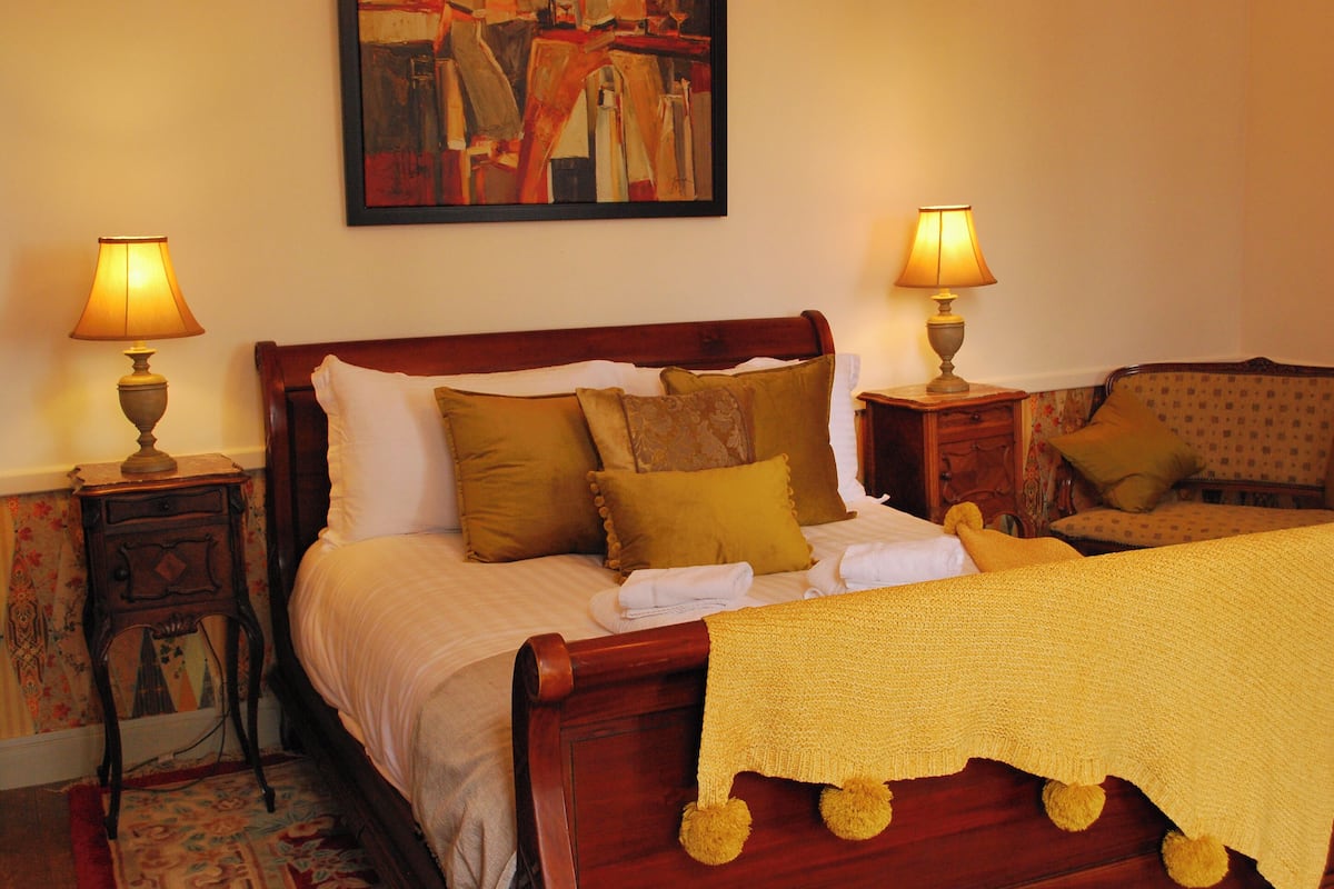 Una cama bien tendida con un edredón amarillo y varios almohadones, flanqueada por dos mesitas de noche con lámparas.