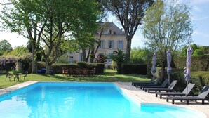 Ein rechteckiger Pool mit Liegestühlen und Sonnenschirmen, umgeben von gepflegten Rasenflächen und Bäumen, mit einem großen Haus im Hintergrund.