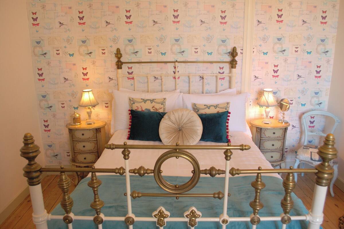 Una cama con estructura metálica, dos mesitas de noche con lámparas y una pared decorada con diseños de mariposas.