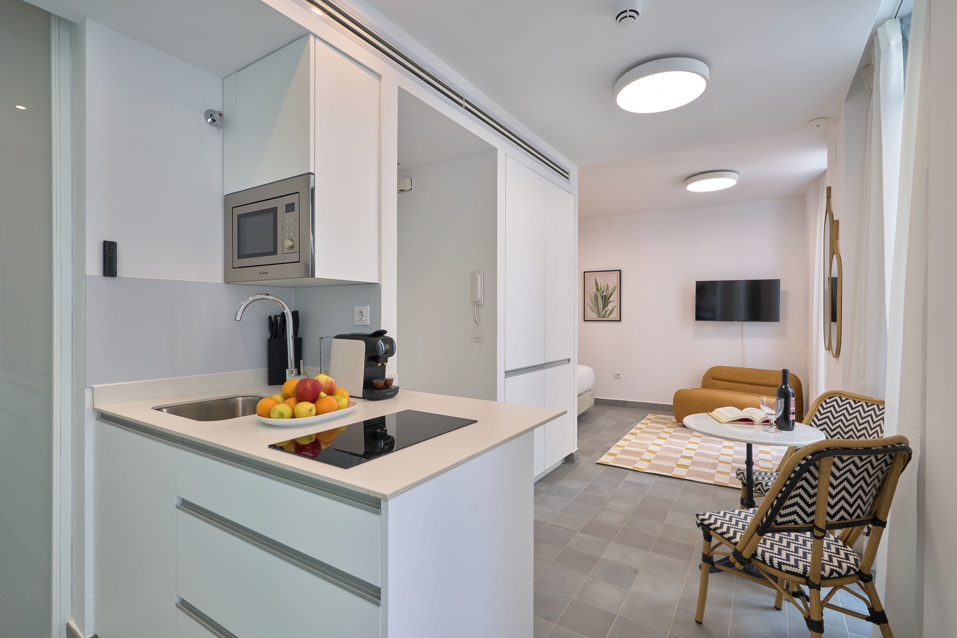 A modern kitchen with a built-in microwave, a coffee machine, and a fruit bowl. There is a dining area with a round table and chairs.