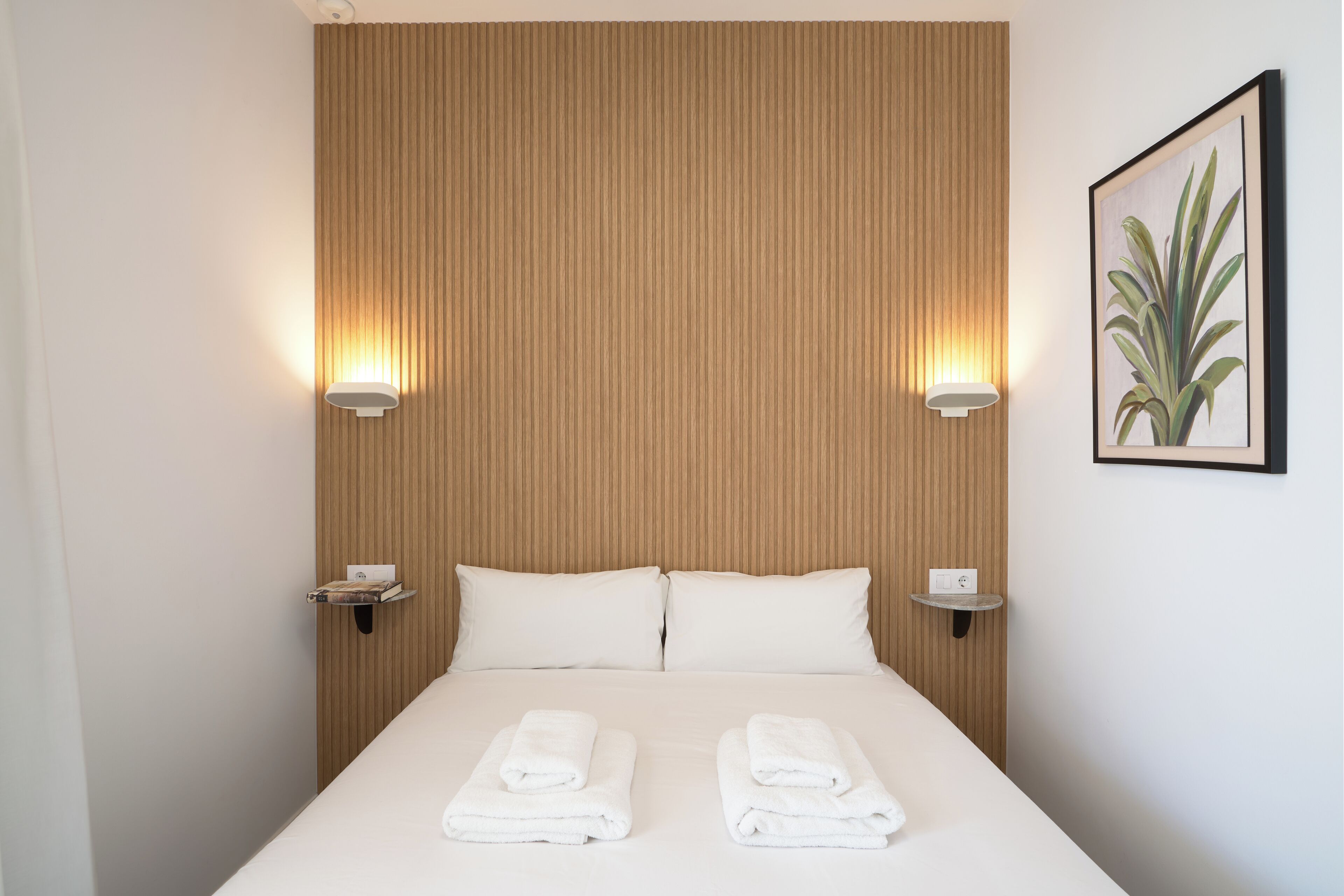 Una habitación de hotel con cabecera de madera, dos lámparas de noche, una planta enmarco en la pared y toallas ordenadas sobre la cama.