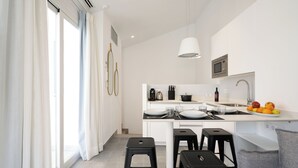 Une cuisine moderne avec des armoires blanches, un four à micro-ondes en acier inoxydable et un panier de fruits sur le comptoir. On y trouve trois tabourets noirs et un canapé gris dans l’espace repas.