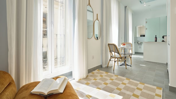 Une chambre d’hôtel moderne avec une grande fenêtre, un tapis à carreaux, un lit en bois, un coin repas avec des chaises et une cuisine à l’arrière-plan.