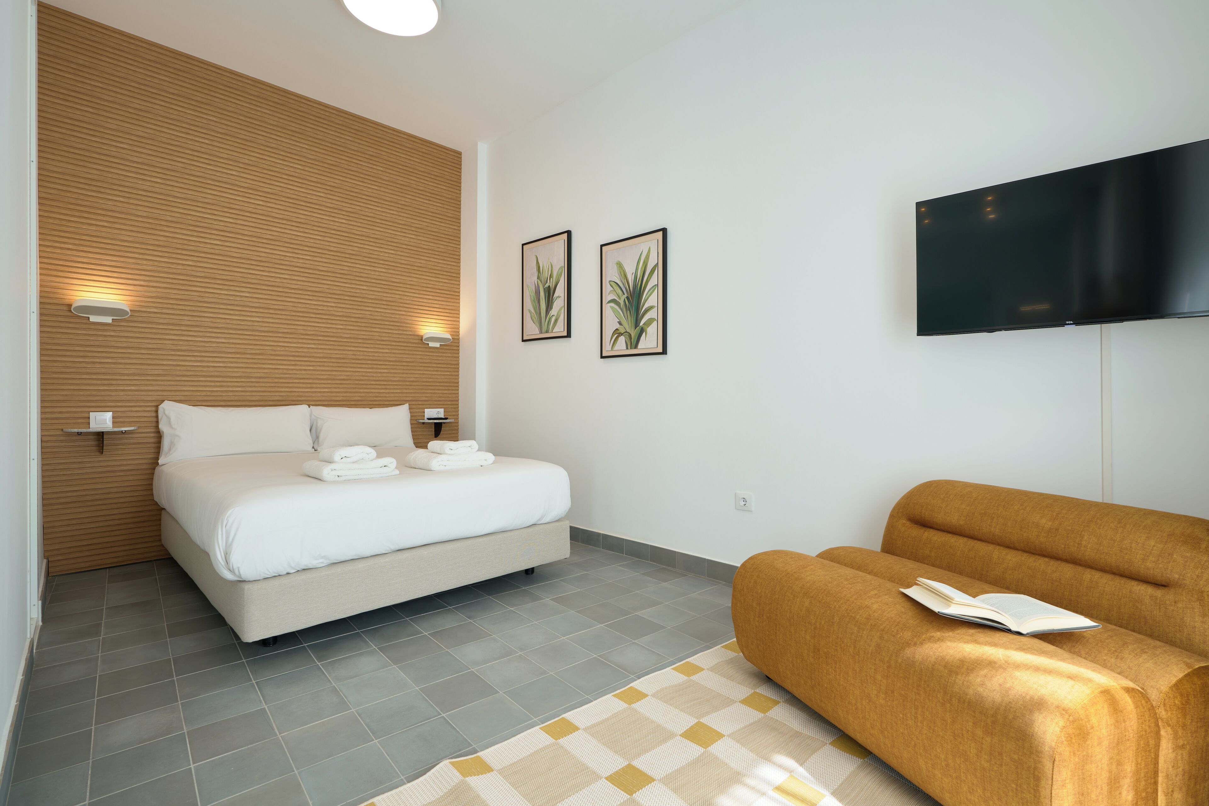 A modern hotel room with a bed, a sofa, a flat-screen TV, and two wall-mounted lights.