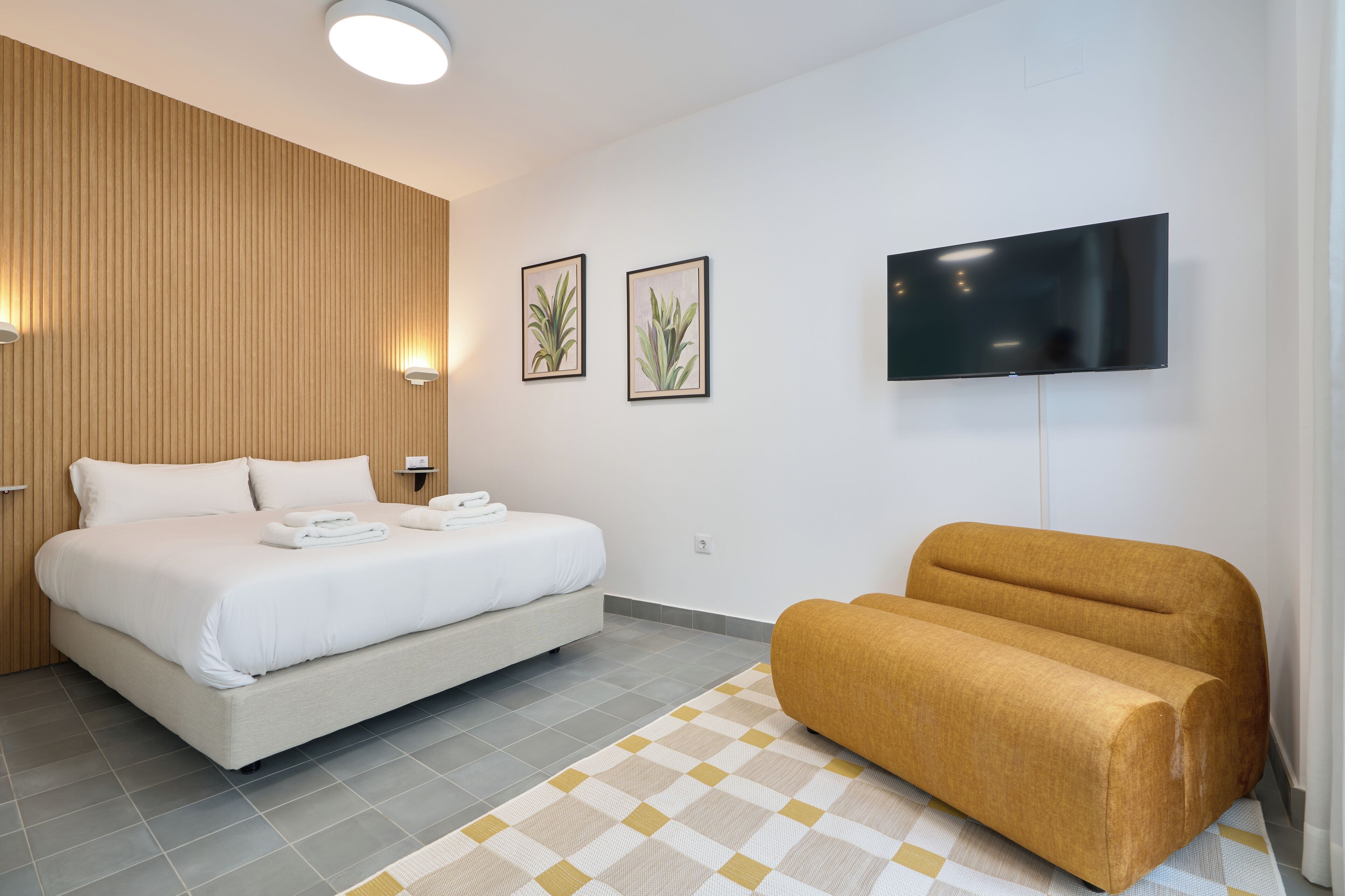 Una habitación de hotel moderna con una cama grande, un sofá, un televisor de pantalla plana y dos plantas en la pared.