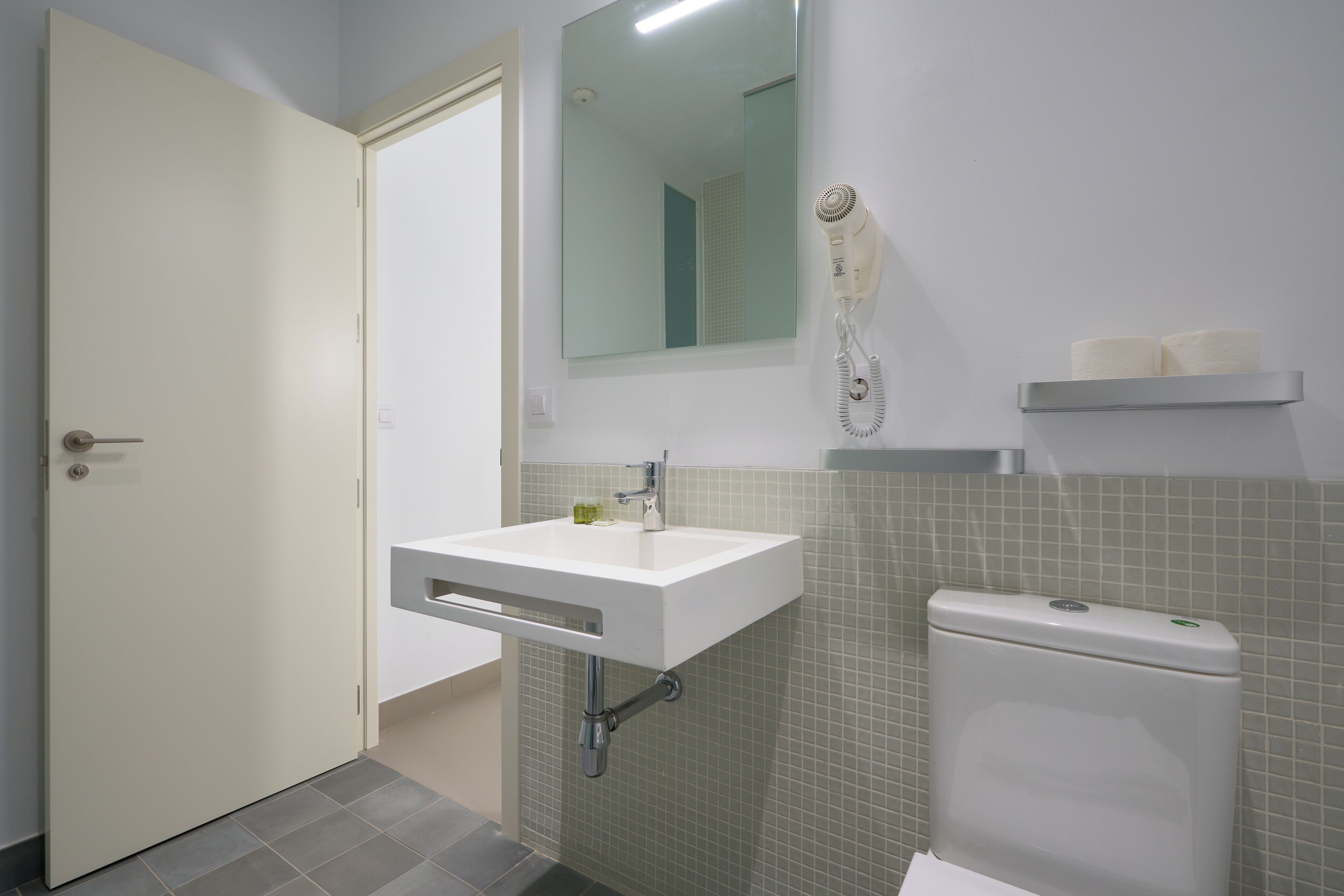 Une salle de bain avec un lavabo blanc, un miroir, un sèche-cheveux et des toilettes.