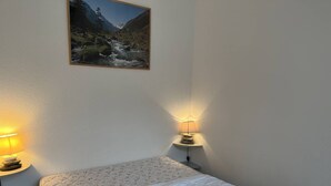 A bedroom with a bed, two bedside lamps, a wall-mounted picture of a mountainous landscape, and a white wall.