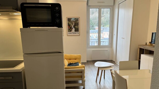 A compact kitchen with a microwave, a small dining table, a yellow chair, and a window with a view of buildings.