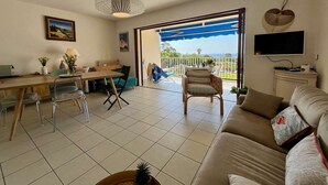 Ein Wohnzimmer mit einer Couch, einem Esstisch, Stühlen, einem Fernseher und Blick ins Freie.