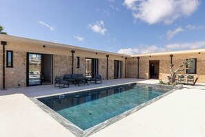 A modern outdoor pool area with a stone facade, outdoor seating, and a clear blue sky.