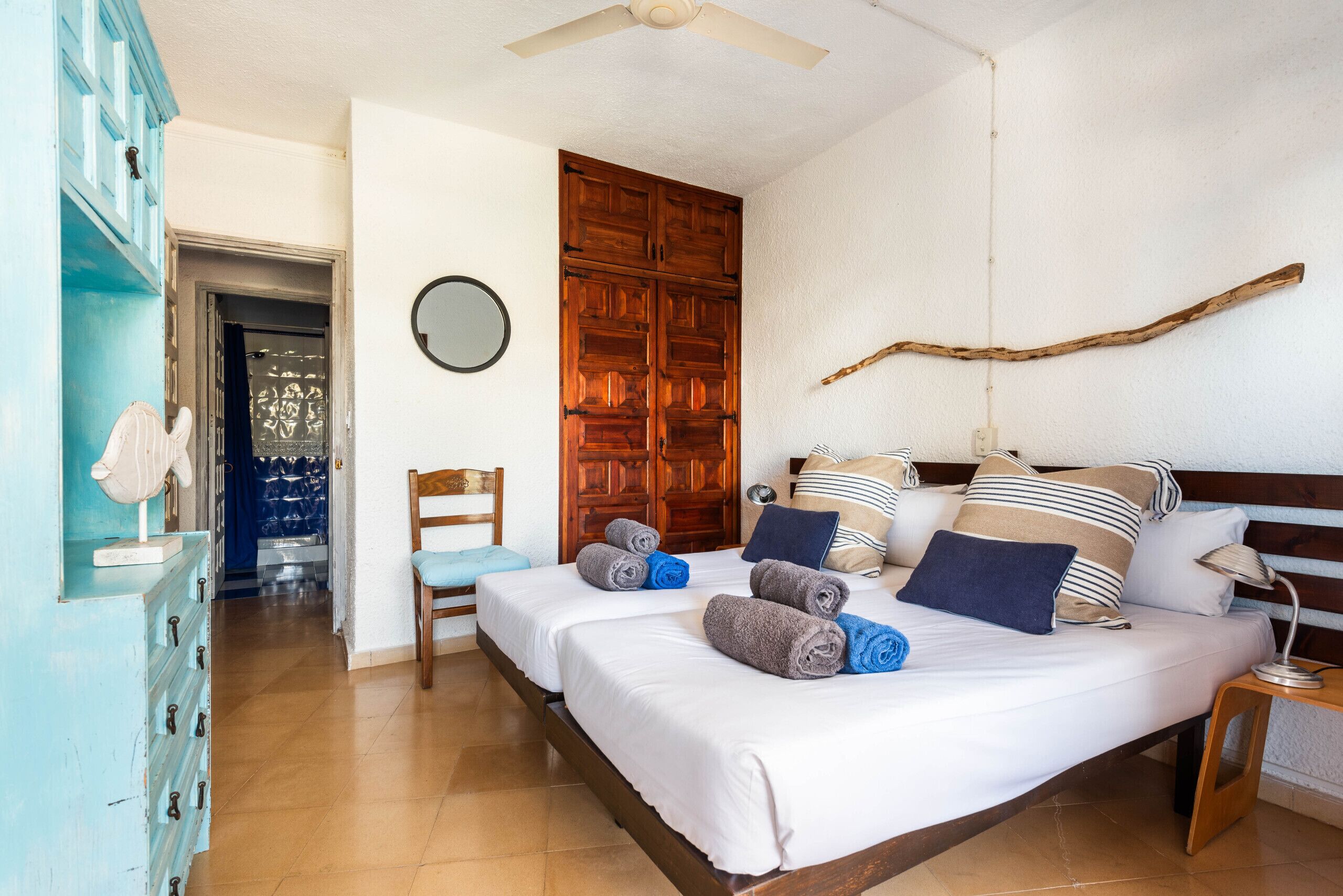 A bedroom with two beds, a wooden wardrobe, a chair, a bedside table with a lamp, and a ceiling fan.