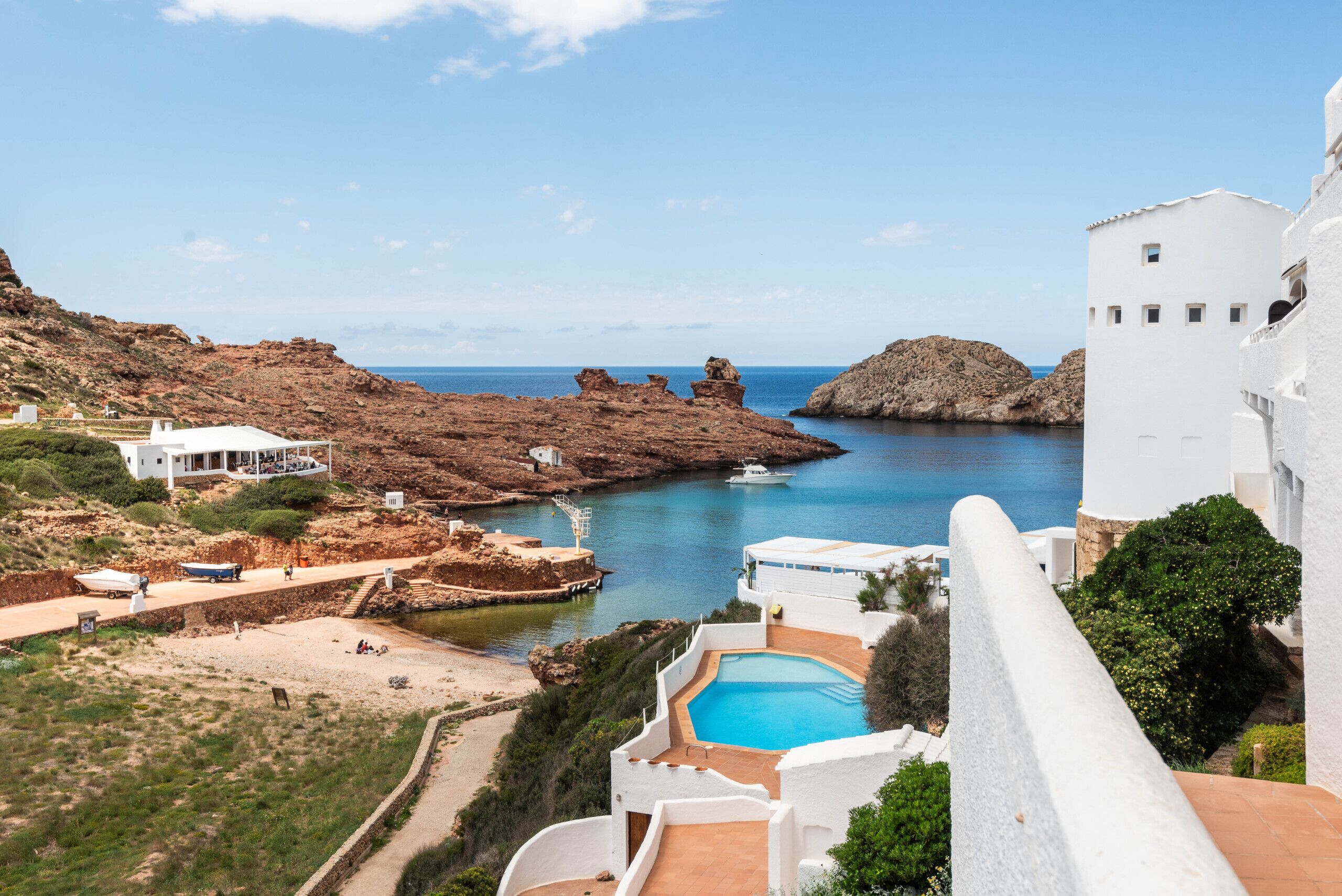 A coastal resort with a swimming pool, white buildings, rocky cliffs, and a clear blue sea.