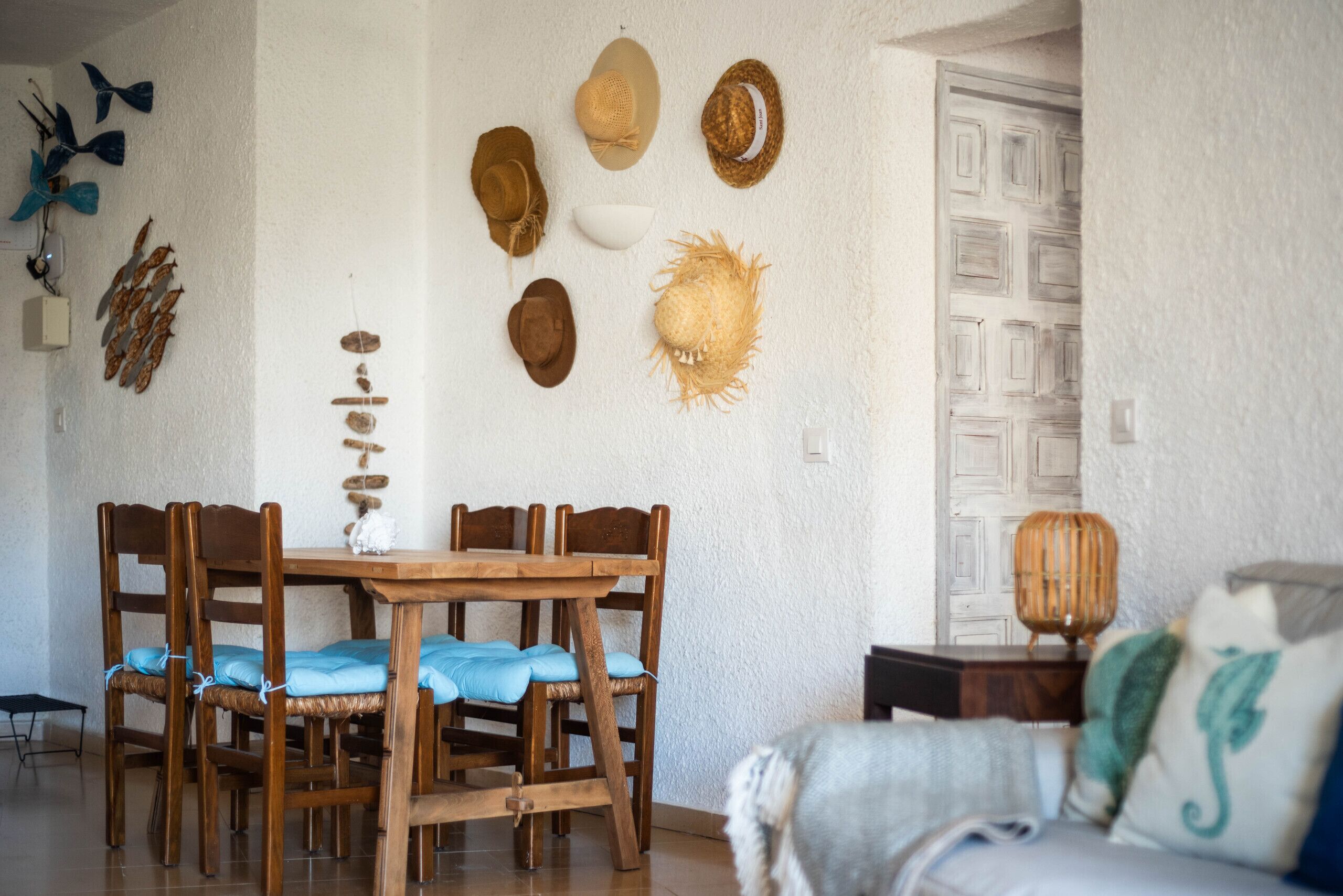 Een zithoek met een houten tafel en stoelen, een witte muur versierd met hoeden, en een bank met een kussen.