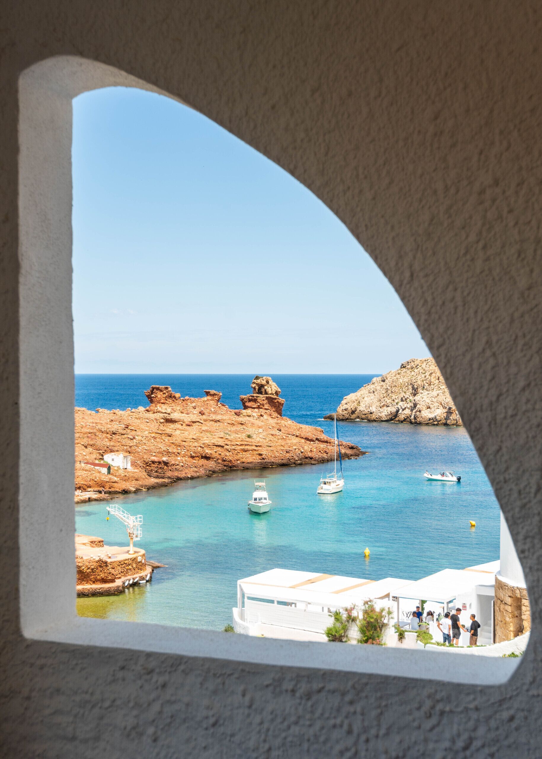 Blick durch ein Fenster auf die Küste, mit felsigem Gelände, blauem Meer, Booten und einem kleinen Gebäude mit Personen.