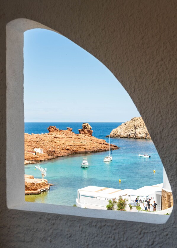 A coastal view through a window, featuring rocky terrain, clear blue sea, boats, and a small building with people.