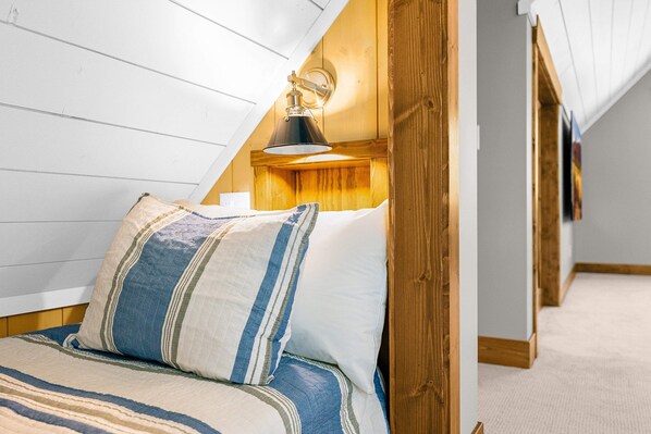 Een slaapkamer met een schuine plafond, een bed met gestreepte beddengoed, en een wandlamp.