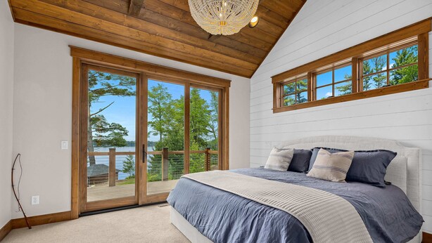 A bedroom with a large bed, wooden ceiling, and a view of trees through a sliding glass door. - NEW! Cross Lake's Finest! (Crosslake)