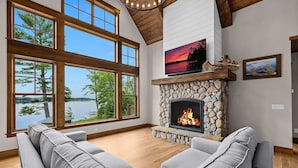 A spacious living room with a stone fireplace, large windows overlooking a lake, and a cozy gray sectional sofa. - NEW! Cross Lake's Finest! (Crosslake)