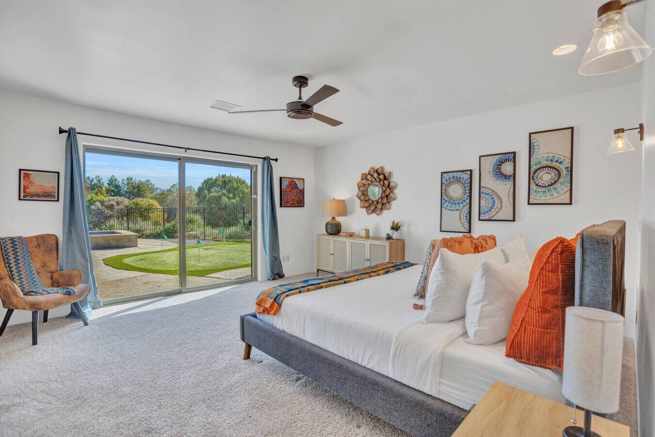 A bedroom with a large sliding glass door leading to a garden, a bed with white bedding, a sofa, and a ceiling fan.