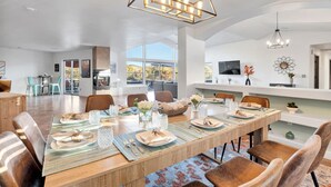 A spacious dining area with a long wooden table set for six, surrounded by comfortable chairs. A modern chandelier hangs above, and a fireplace is visible in the background. - Serene Retreat - Heated Pool, Spa, Sauna, Grass Pickleball (Sedona)