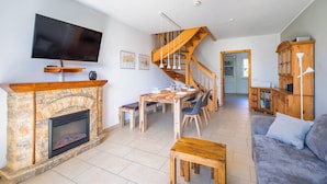 Un salon avec une cheminée, un téléviseur fixé au mur, un escalier en bois et un coin repas avec une table et des chaises.